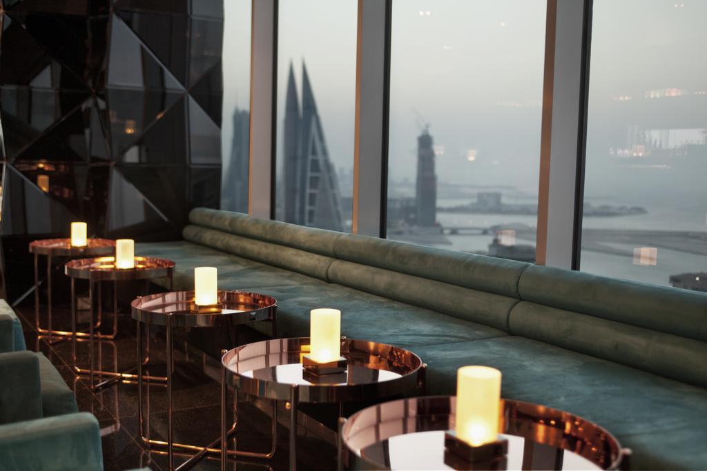 The Domain Bahrain Hotel And Spa - Adults Friendly 16 Years Plus Manama Eksteriør billede The photo depicts a stylish lounge area with a modern aesthetic. There are several circular tables made of metallic materials, each topped with a small glowing candle. A long, plush seating area in a teal color runs along the window, offering a view 